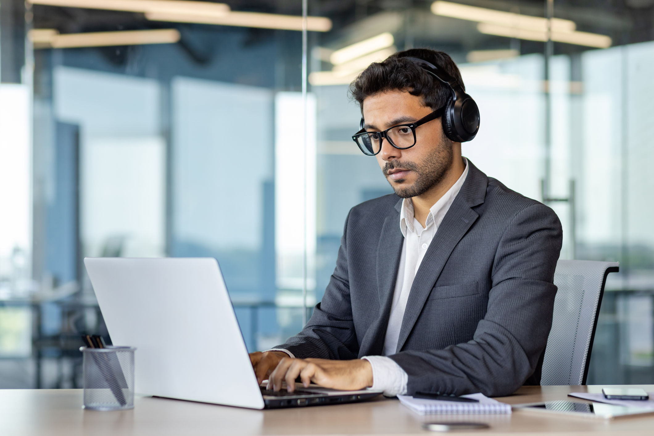Serious man working inside office with laptop, concentrated and thinking businessman boss in business suit using headphones listening to music and educational audio course and book